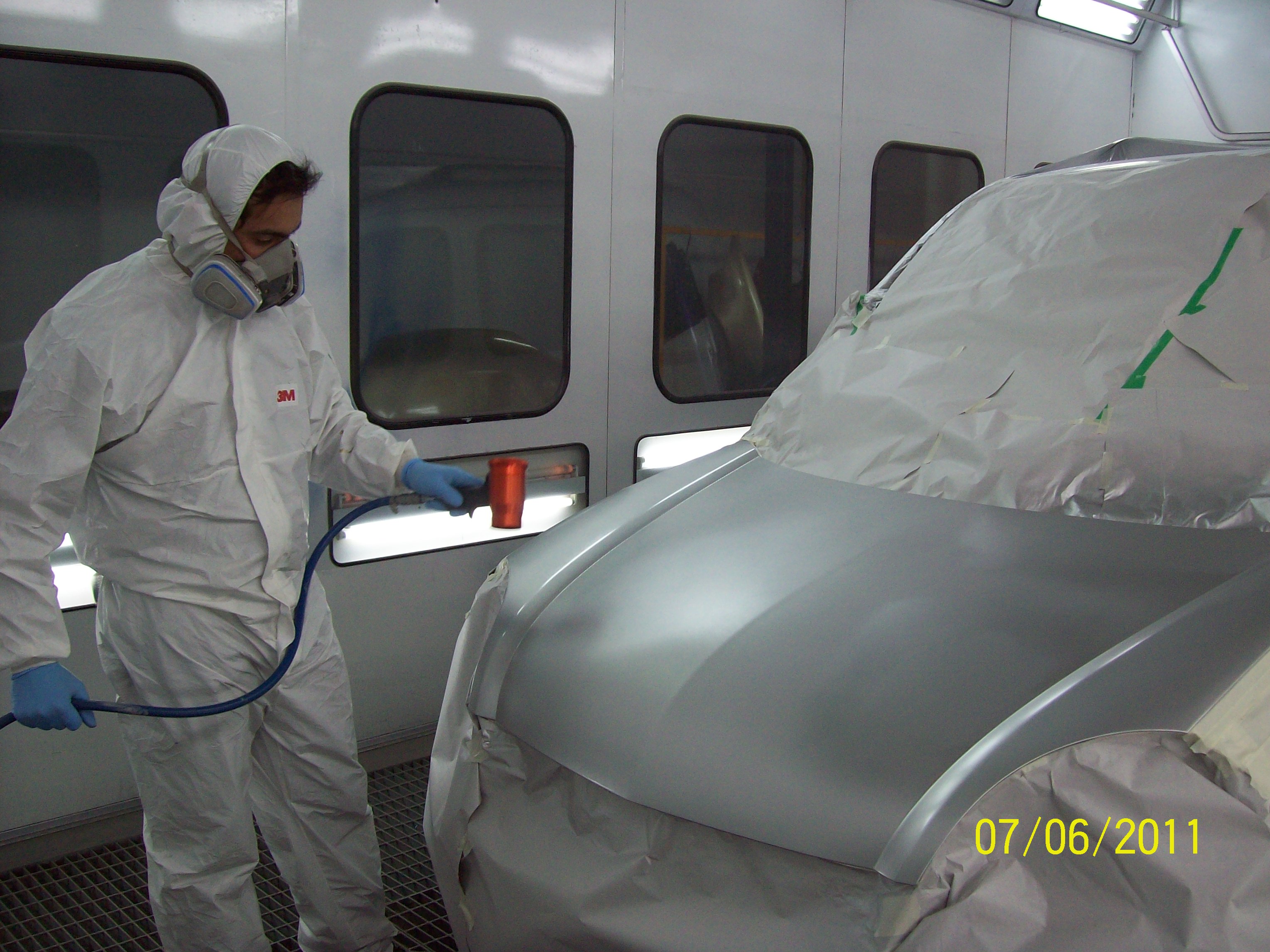 Persona con mameluco y accesorios de protección pintando un auto en una cabina de pintura.